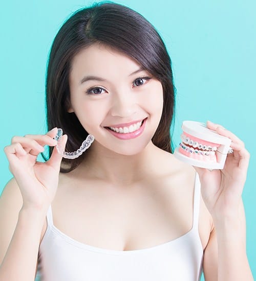 Woman holding up a clear aligner and model smile with traditional braces